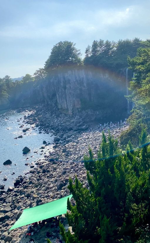 The image shows a scenic view of Jeongbang Waterfall in Jeju Island, South Korea. A faint rainbow arches over the cliff where the waterfall descends into the rocky shoreline below. The area is surrounded by lush green trees, with a few people visible near the water's edge. In the foreground, a green canopy provides shade to visitors enjoying the natural beauty of the scene. The overall atmosphere is peaceful, enhanced by the natural rainbow formation.