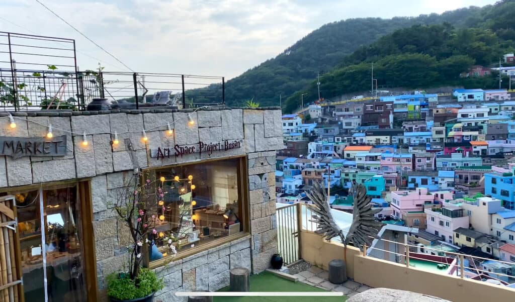 View from a café at Gamcheon Culture Village in Busan, South Korea, showcasing a vibrant tapestry of colorful houses densely packed on a hillside, with lush greenery in the background and traditional Korean café décor in the foreground