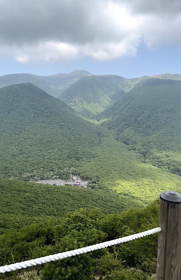 Lush green mountains with a cloudy sky, a parking lot visible at the base, and a wooden railing in the foreground.