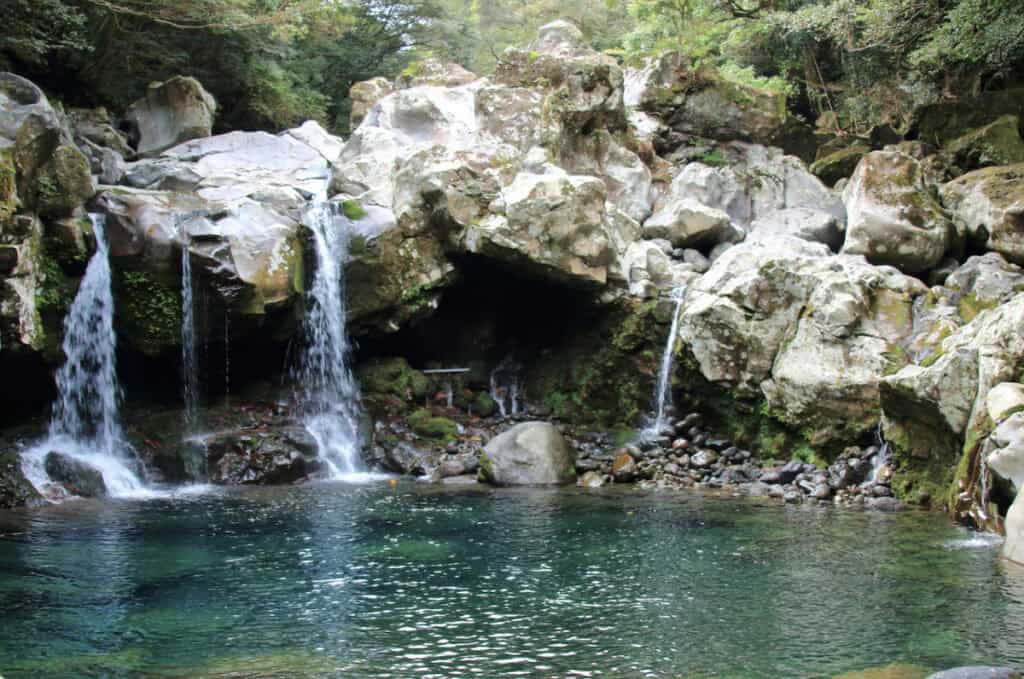 This image features a serene scene of Wonang Waterfall in Jeju Island, showcasing a crystal-clear pond fed by a twin set of waterfalls cascading over a rugged, moss-covered rock face, with lush greenery surrounding the tranquil pool.