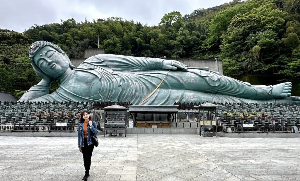 A person stands in front of the massive Reclining Buddha statue at Nanzoin Temple, surrounded by lush green trees.