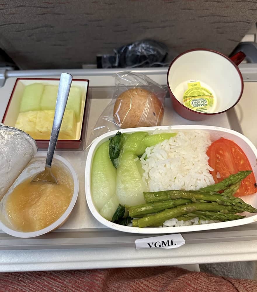 Vegan breakfast tray on a flight, featuring steamed rice, asparagus, bok choy, tomato slices, and sides of fruit and bread.