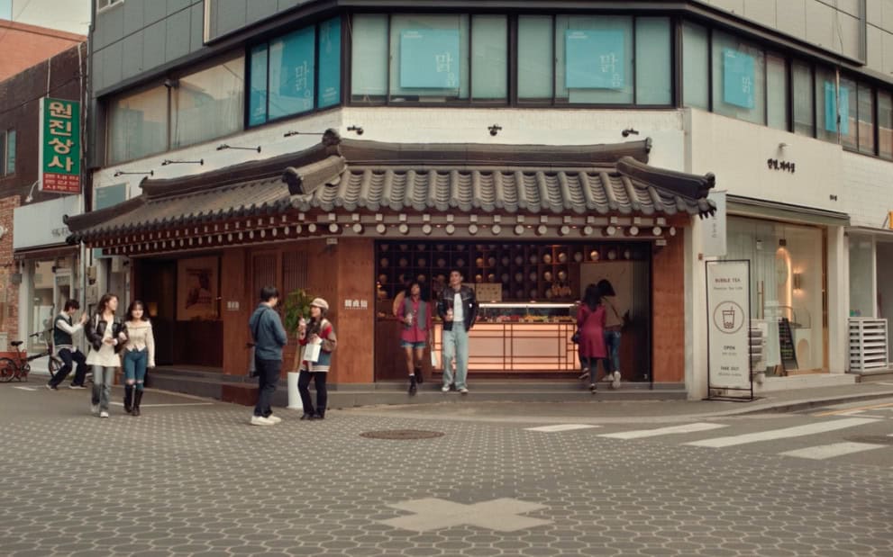 A modern café and boutique area in Seongsu, Seoul, with characters walking past a hanok-style bubble tea shop. The scene highlights the blending of traditional and contemporary styles.