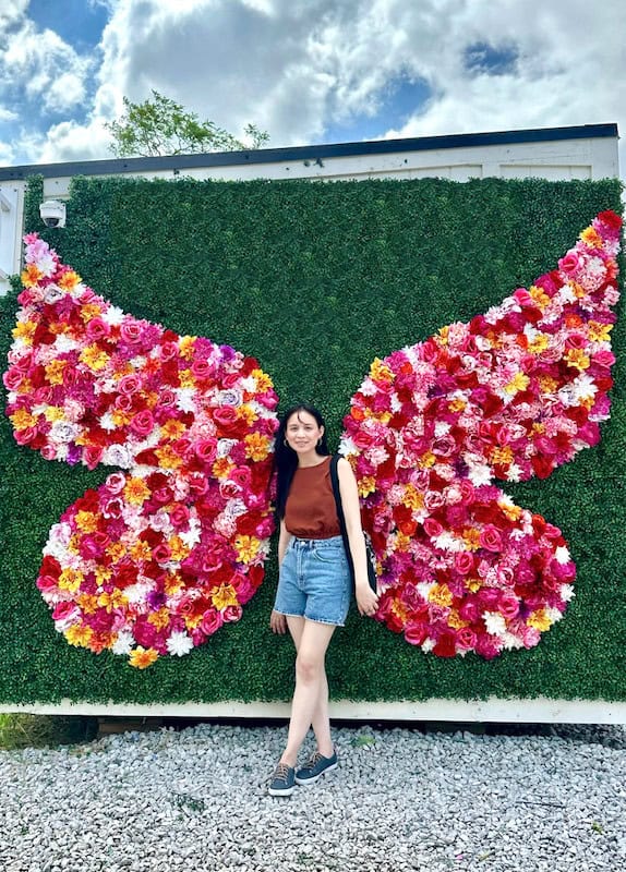A woman stands against a green hedge wall with oversized butterfly wings made from pink red yellow and white flowers spreading behind her.